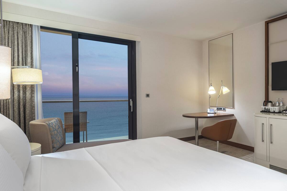 A hotel room with a bed, a desk, a chair, a TV, and a view of the ocean.