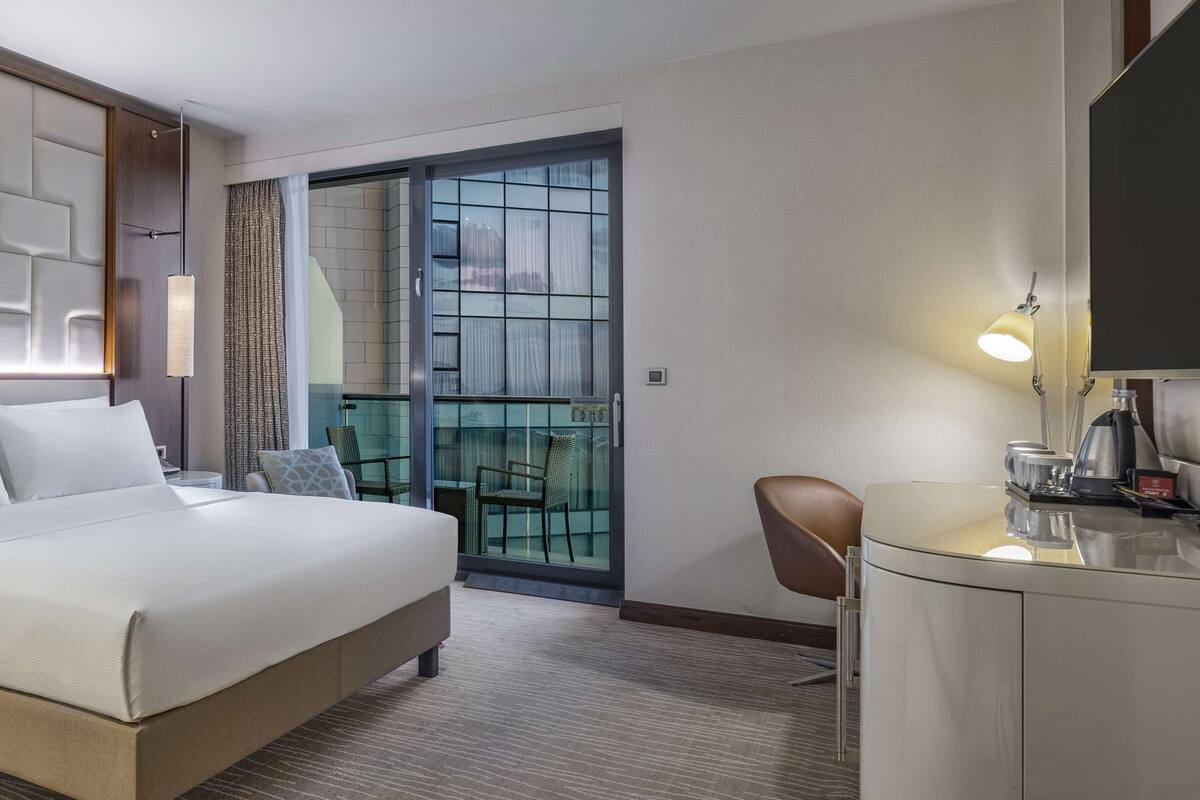 A hotel room with a bed, bedside tables, a desk with a lamp, a chair, and a view of the outside through a glass door.