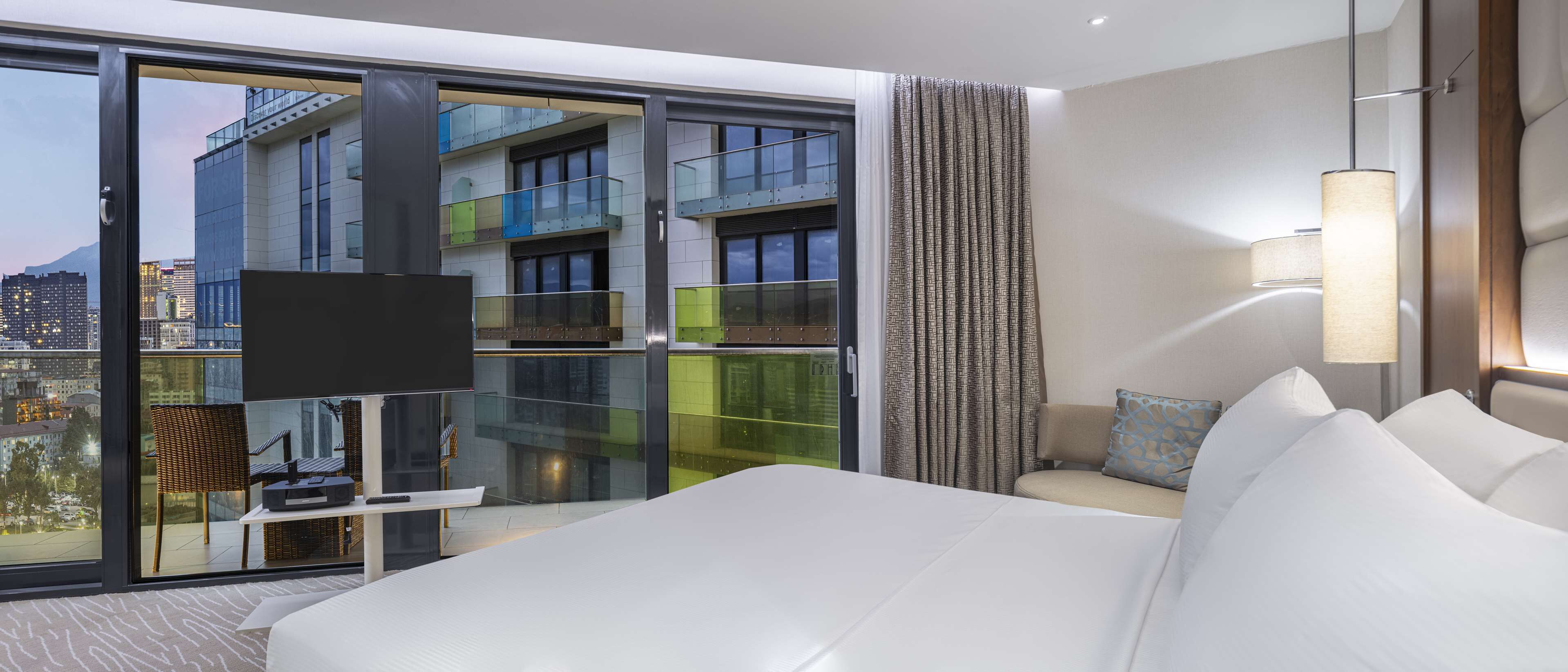 A modern hotel room with a large bed, a TV, a sofa, and a view of a cityscape through floor-to-ceiling windows.