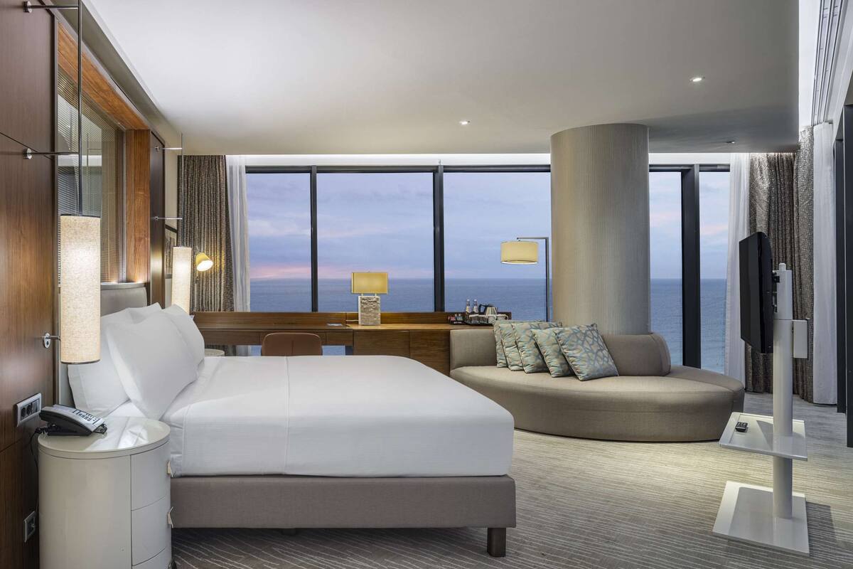 A modern hotel room with a large bed, a desk, a sofa, and a view of the ocean.