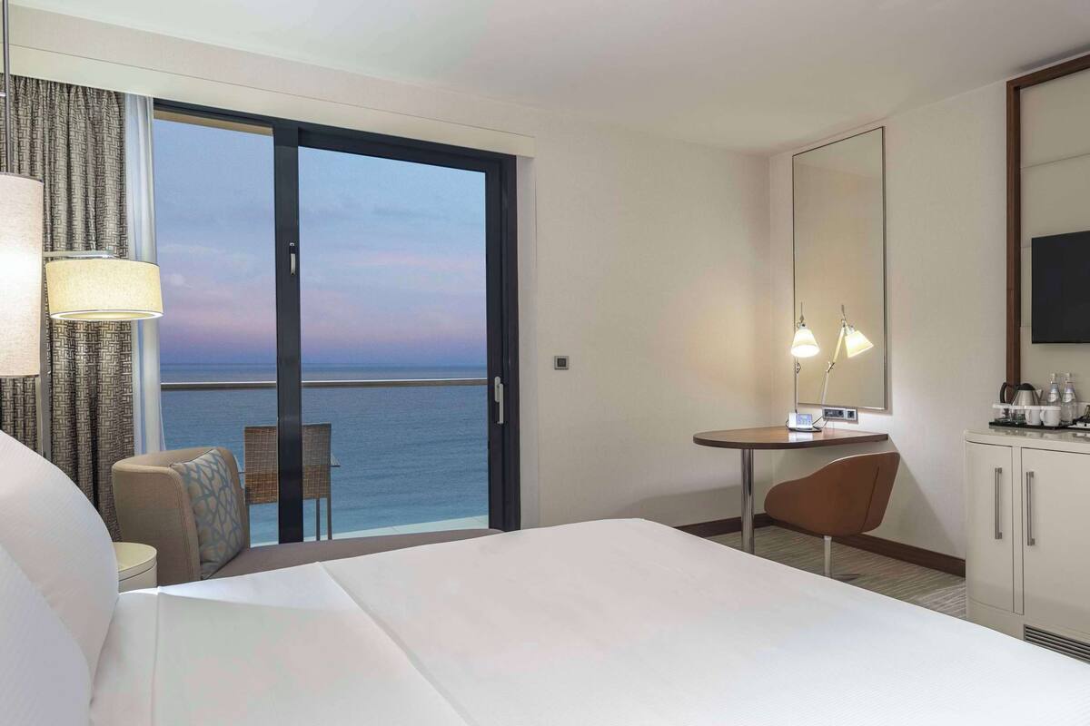 A hotel room with a bed, a desk, a chair, a TV, and a view of the ocean.