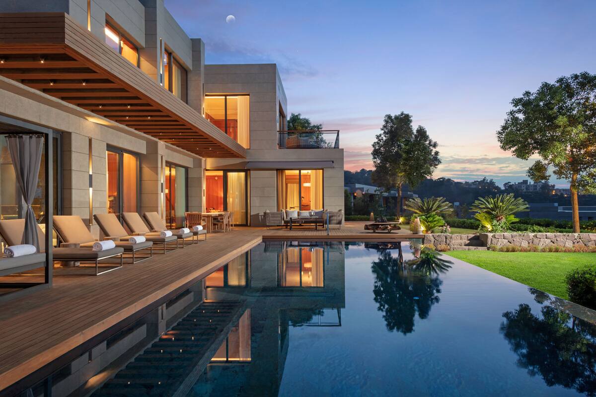 A modern house with a swimming pool, lounge chairs, and a view of the city at dusk.