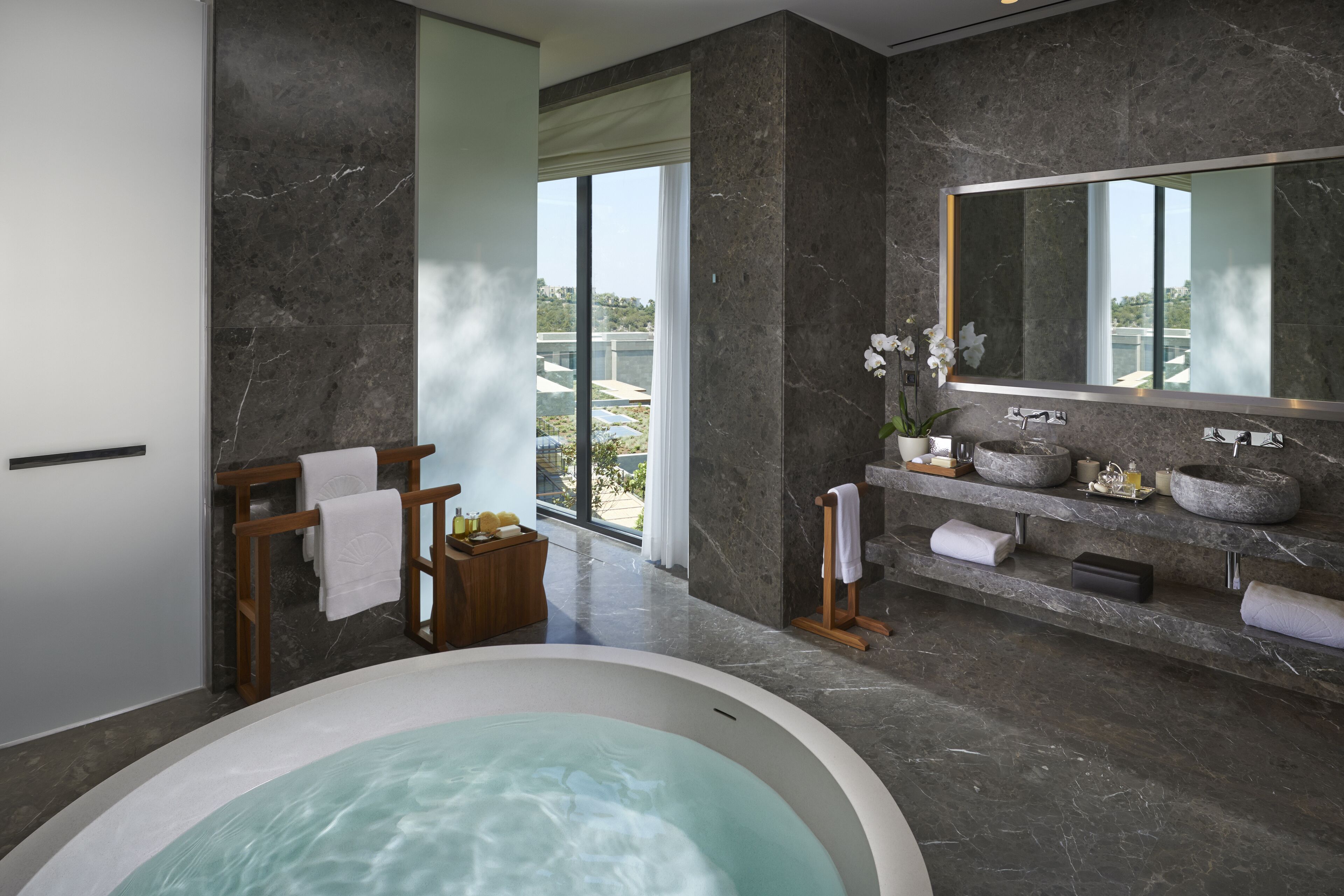 A modern bathroom with a large bathtub, marble walls, a wooden vanity with stone sinks, and a large mirror.
