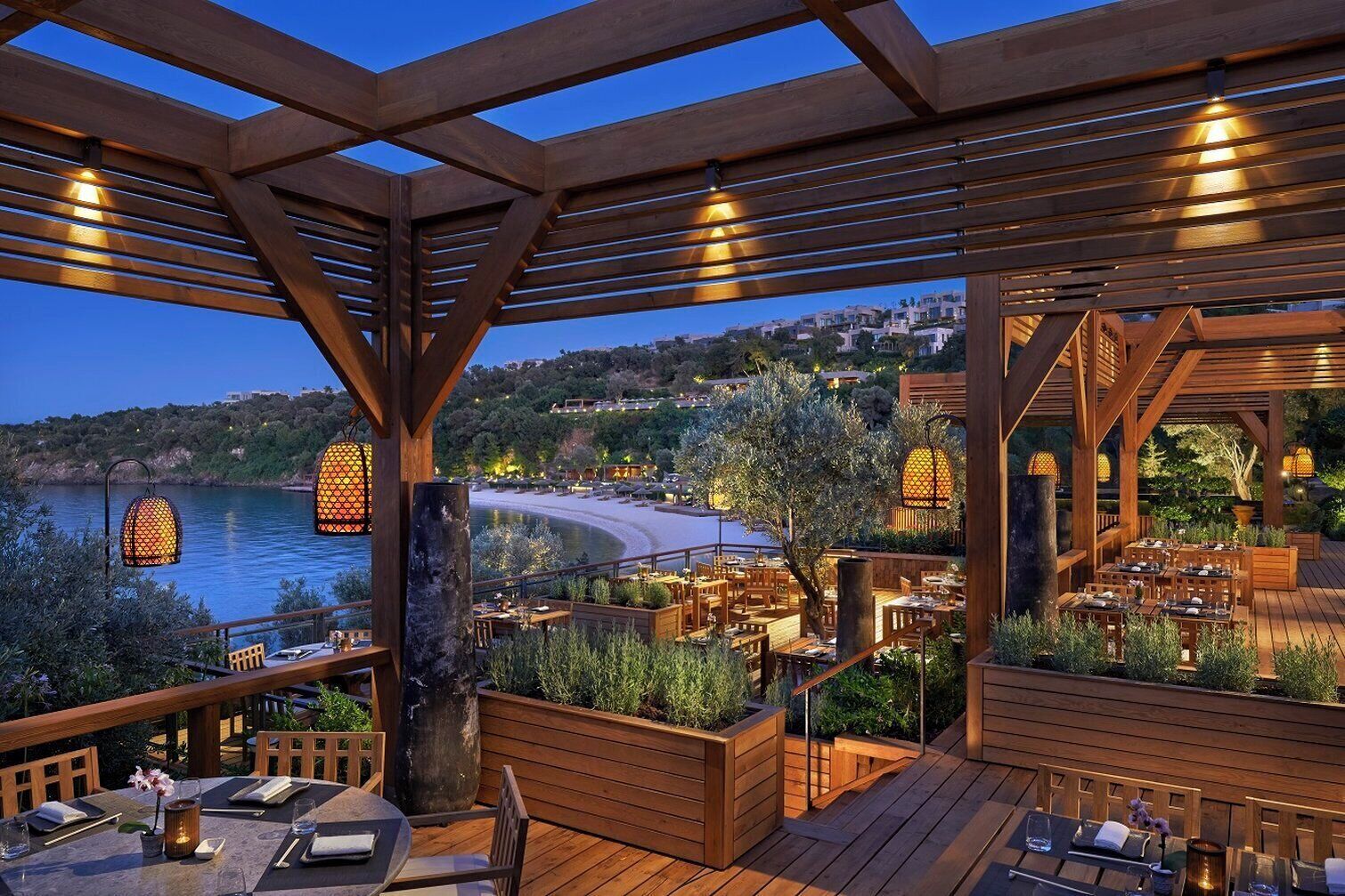 Un espacio de comedor al aire libre con pérgolas de madera, mesas y sillas, con vista a una playa y colinas.