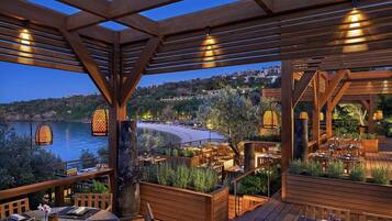 Un espacio de comedor al aire libre con pérgolas de madera, mesas y sillas, con vista a una playa y colinas.