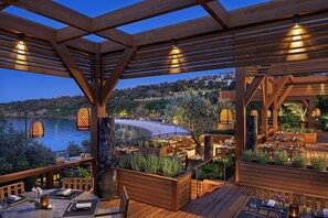 An outdoor dining area with wooden pergolas, tables, and chairs, overlooking a beach and hills. - Mandarin Oriental, Bodrum (Bodrum)