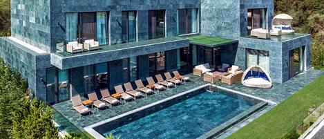 Un edificio moderno de dos pisos con piscina, sillas de descanso y una terraza con paredes de vidrio.