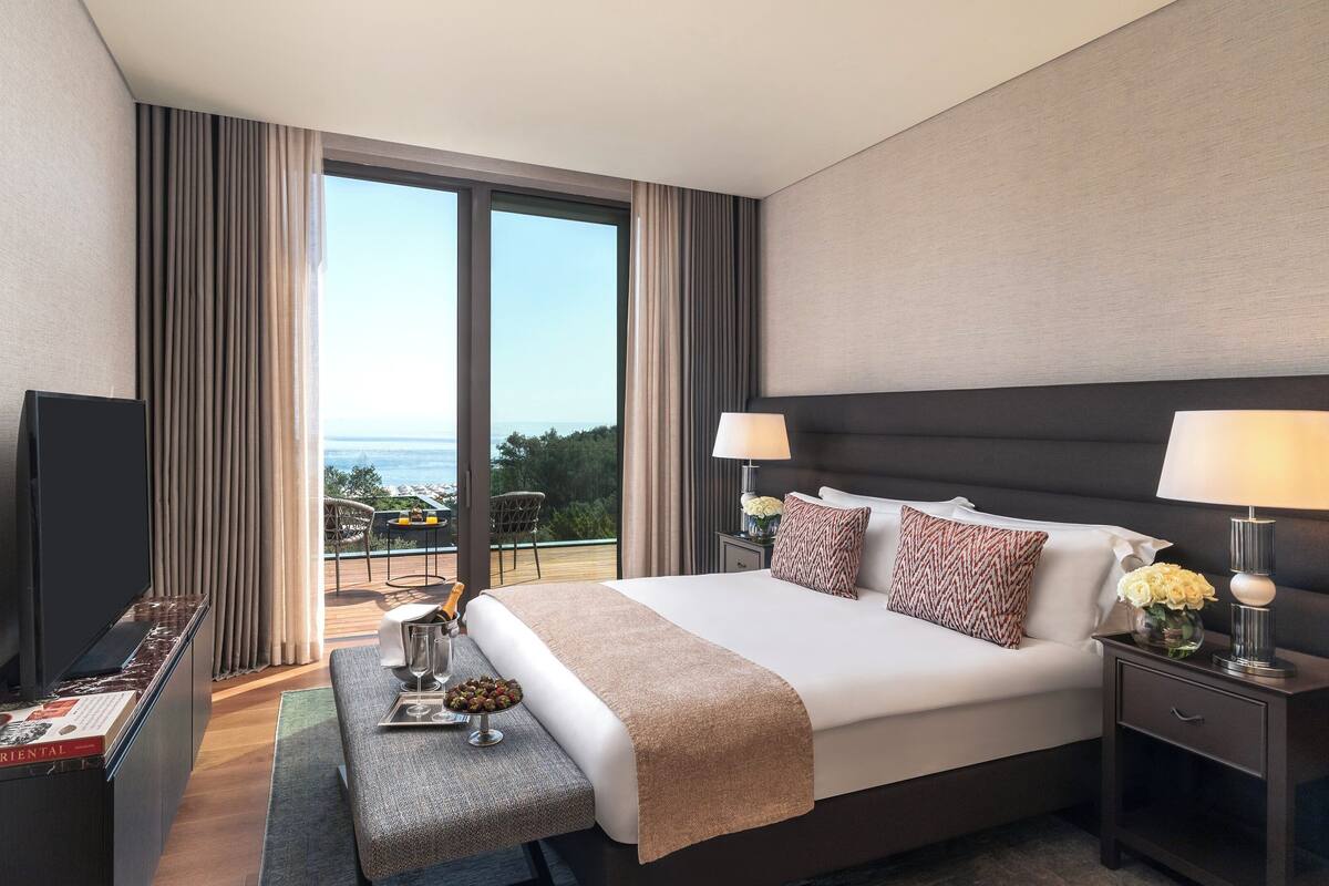 A hotel room with a large bed, two bedside lamps, a TV, a small table with a bottle and glasses, and a view of the ocean.