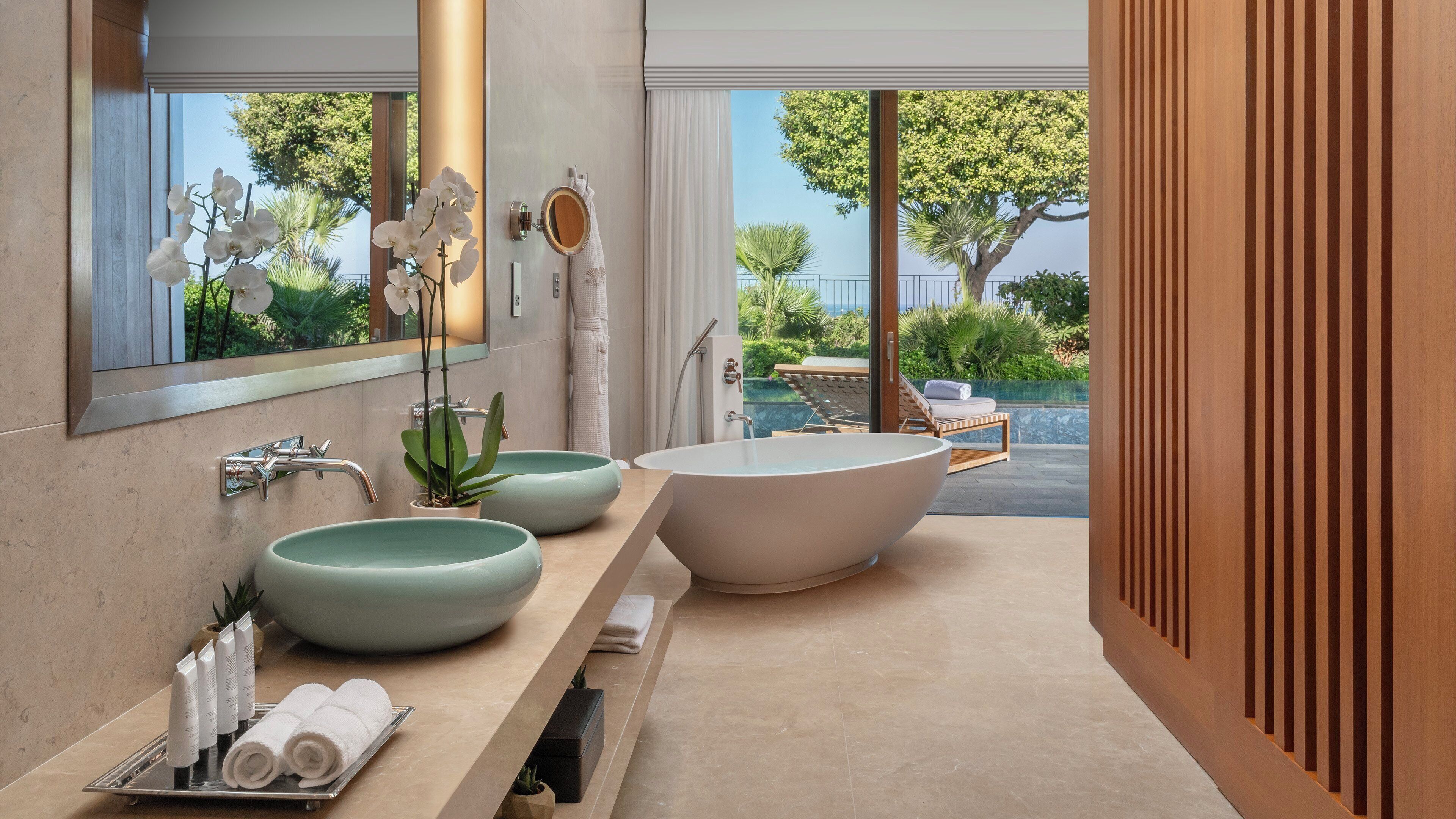 Une salle de bain moderne avec une grande baignoire sur pieds, deux vasques rondes, un miroir et une vue sur une piscine et la verdure environnante.