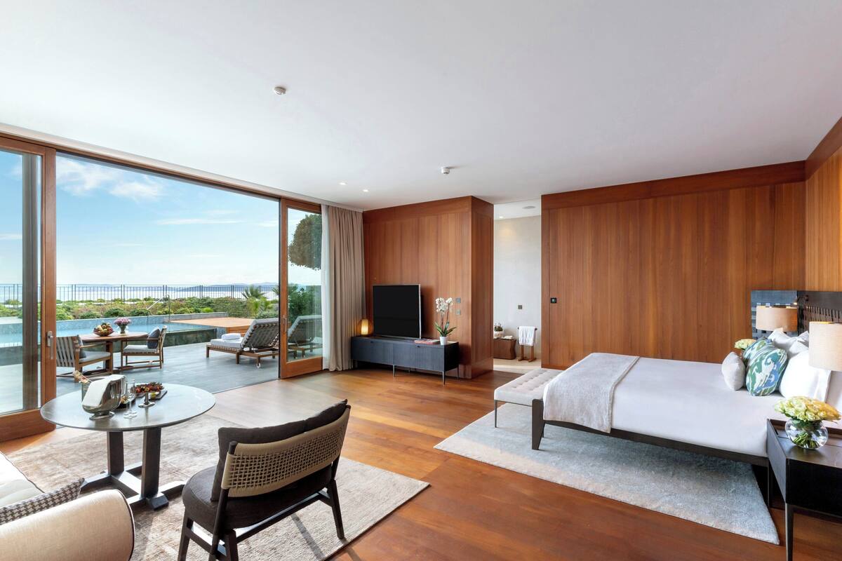 A modern hotel room with a large balcony, a bed, a TV, and a dining area with a table and chairs.