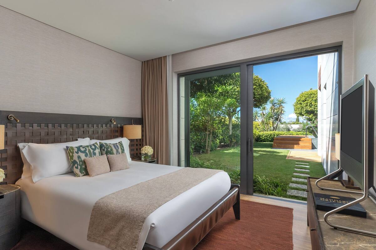 A modern hotel room with a large bed, a TV, and a view of a garden through a sliding glass door.