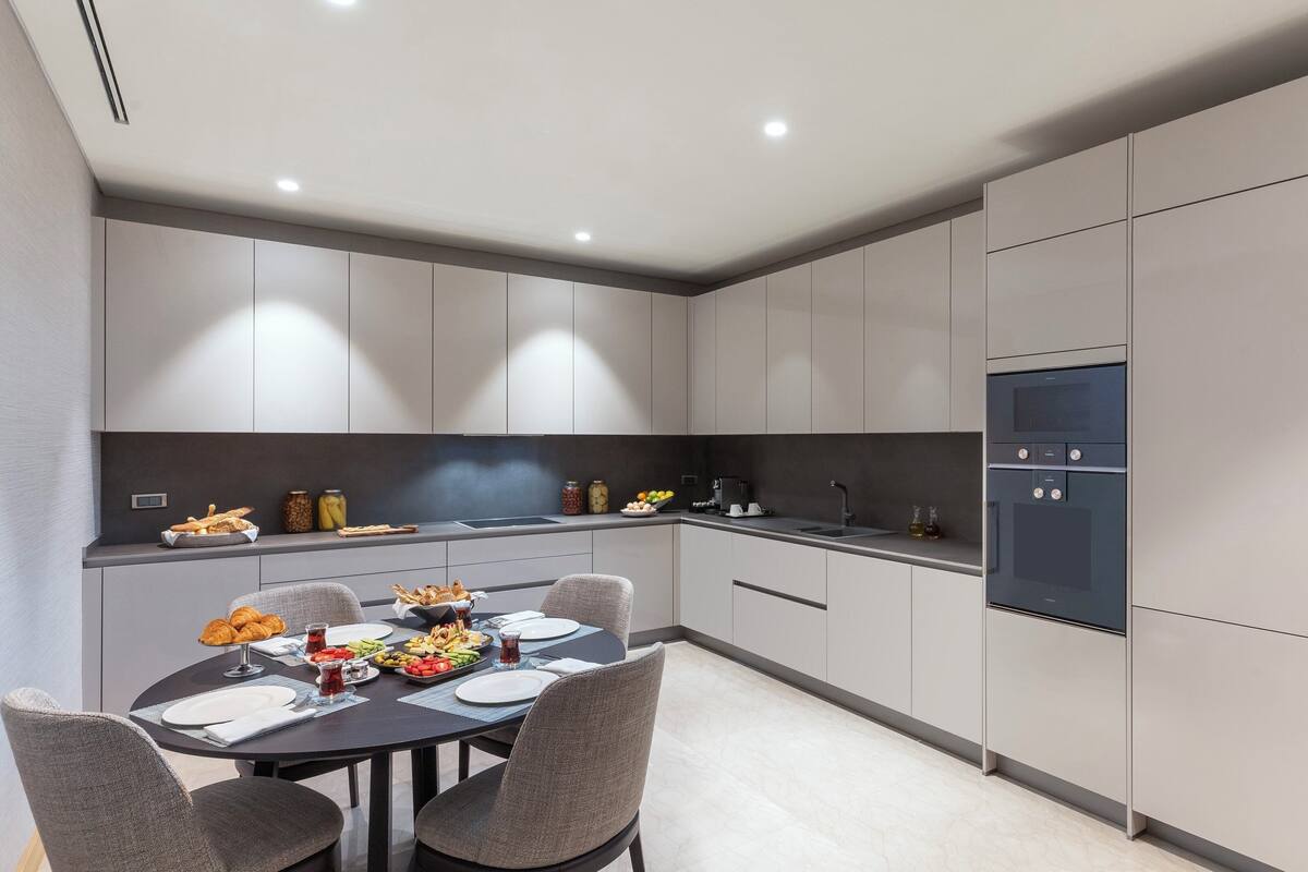 A modern kitchen with grey cabinets, a black countertop, and a built-in oven. There is a dining table with chairs and a variety of food items on it.