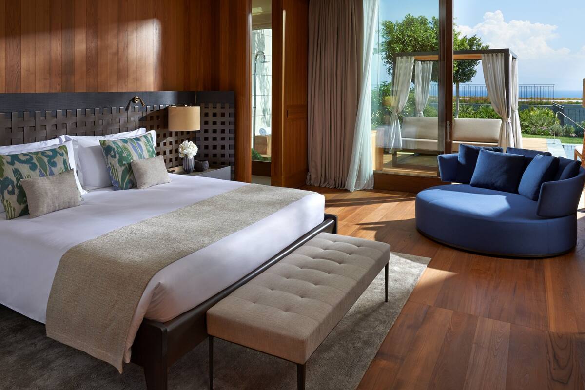 A hotel room with a large bed, a sofa, a bench, and a view of the outdoors.