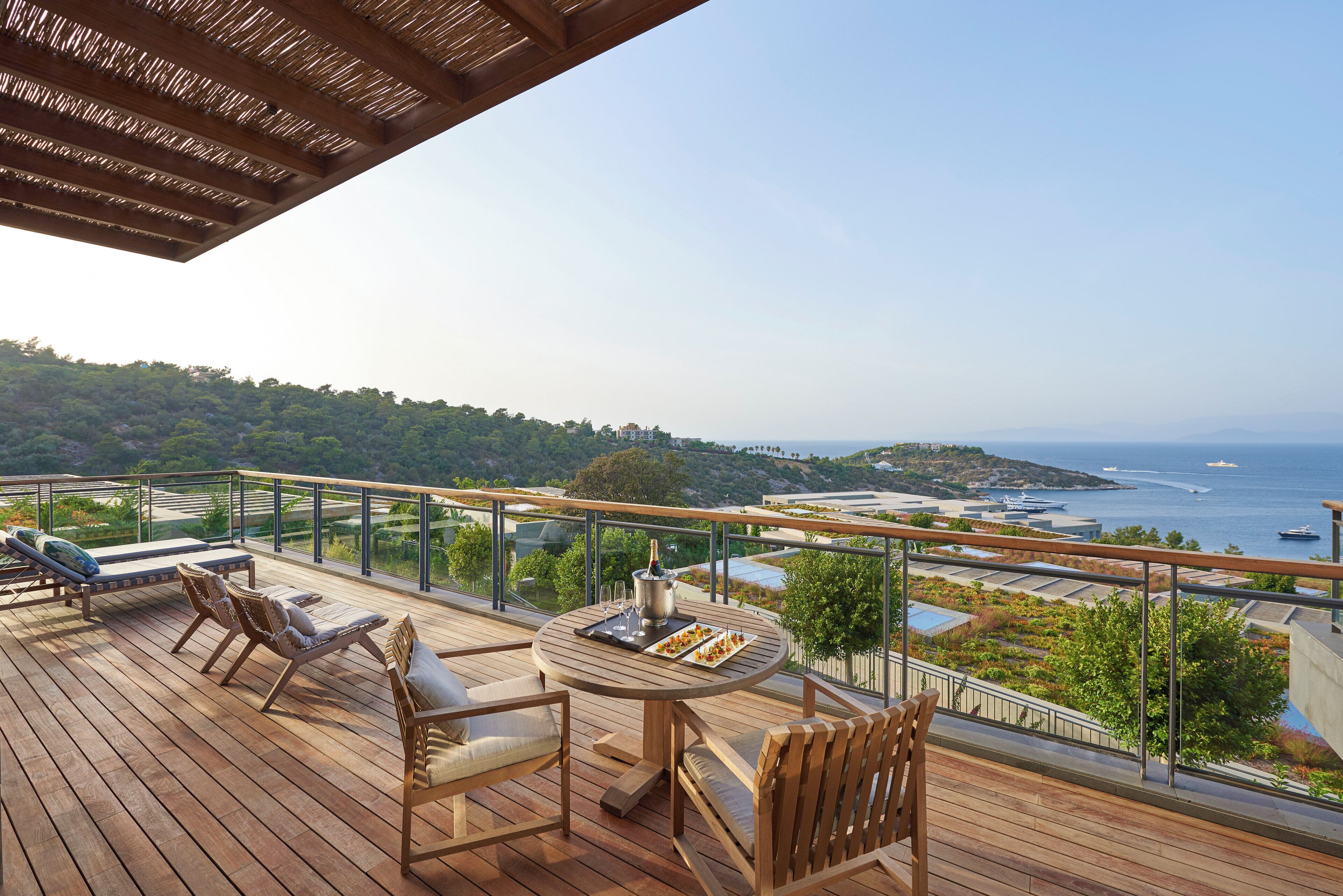 A wooden deck with lounge chairs and a table, overlooking a coastal area with boats and greenery.