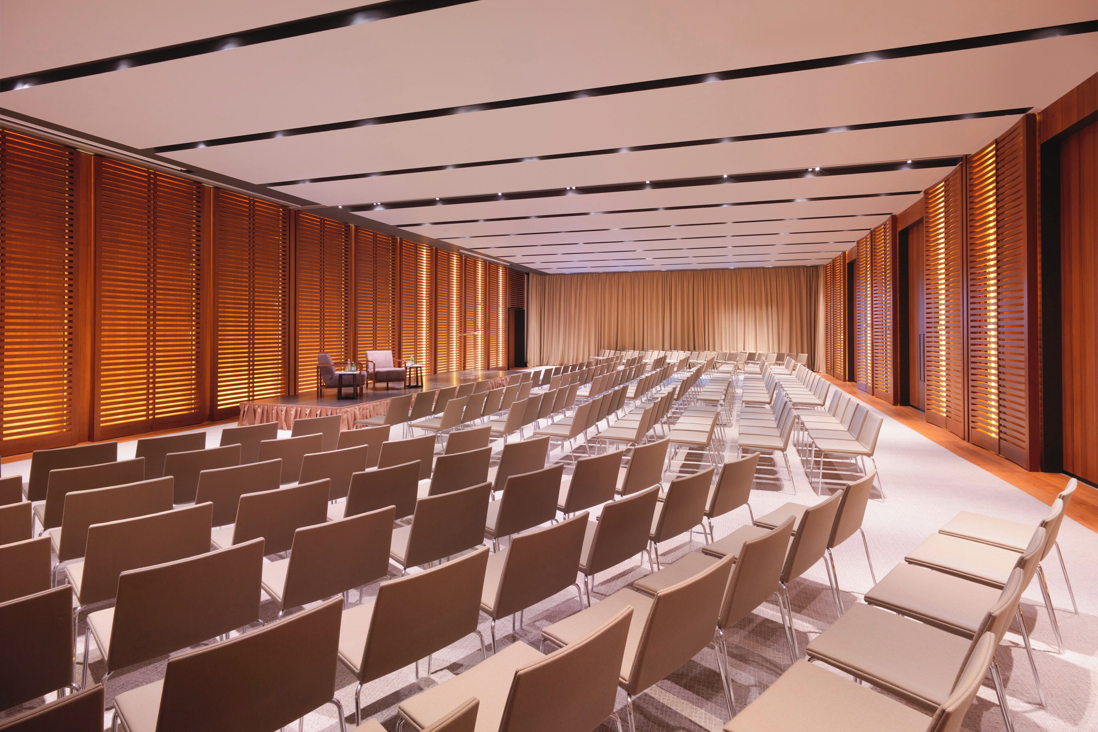A conference room with rows of chairs, a stage, and wooden paneling on the walls.