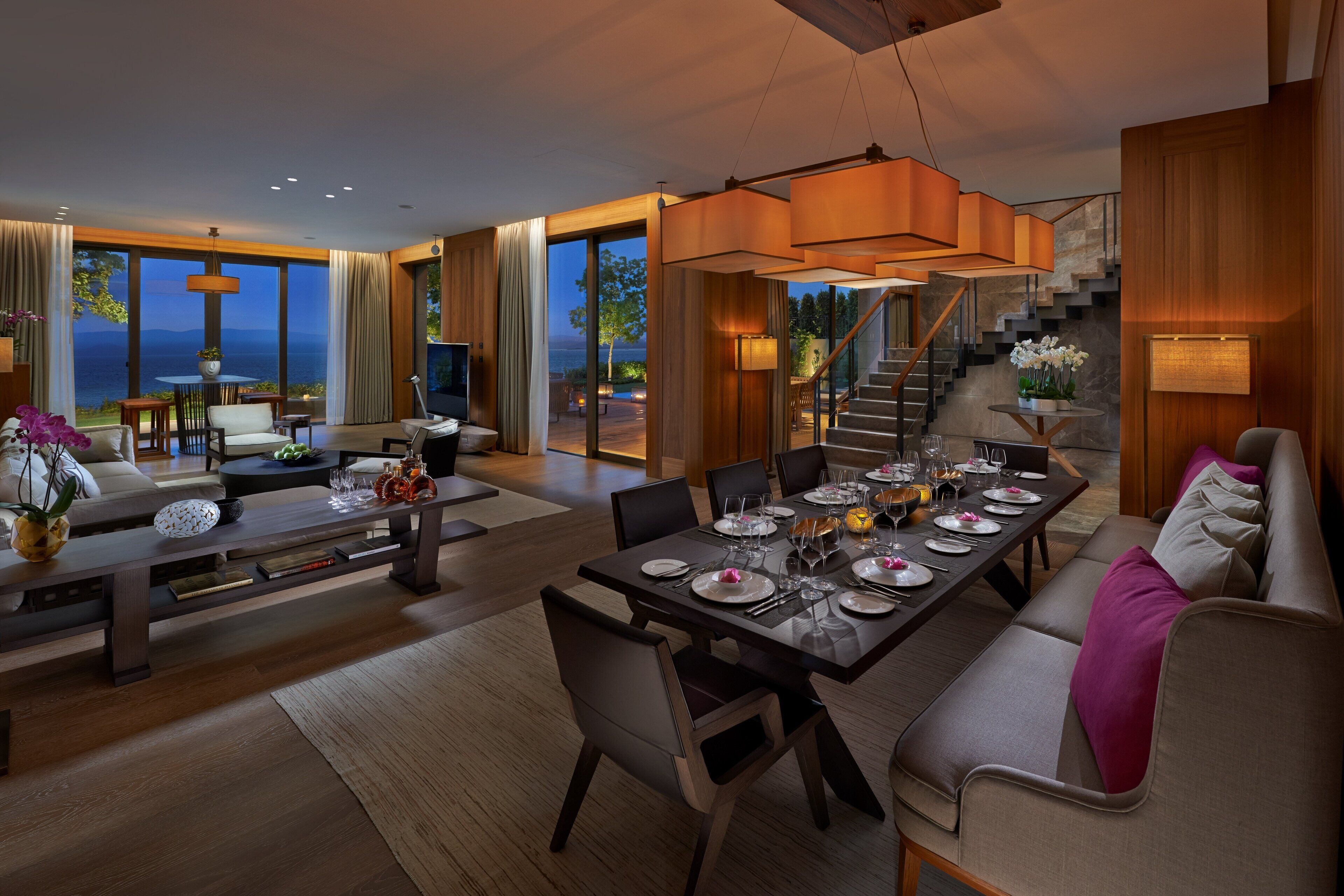 Un salón amplio con una gran mesa de comedor dispuesta para una comida, un sofá con cojines, y una escalera que lleva a un espacio exterior con vistas al mar.