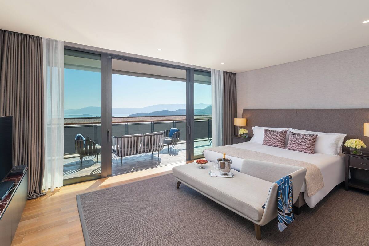 A modern hotel room with a large bed, a TV, a balcony with a view of the mountains, and a small table with a bottle of champagne.
