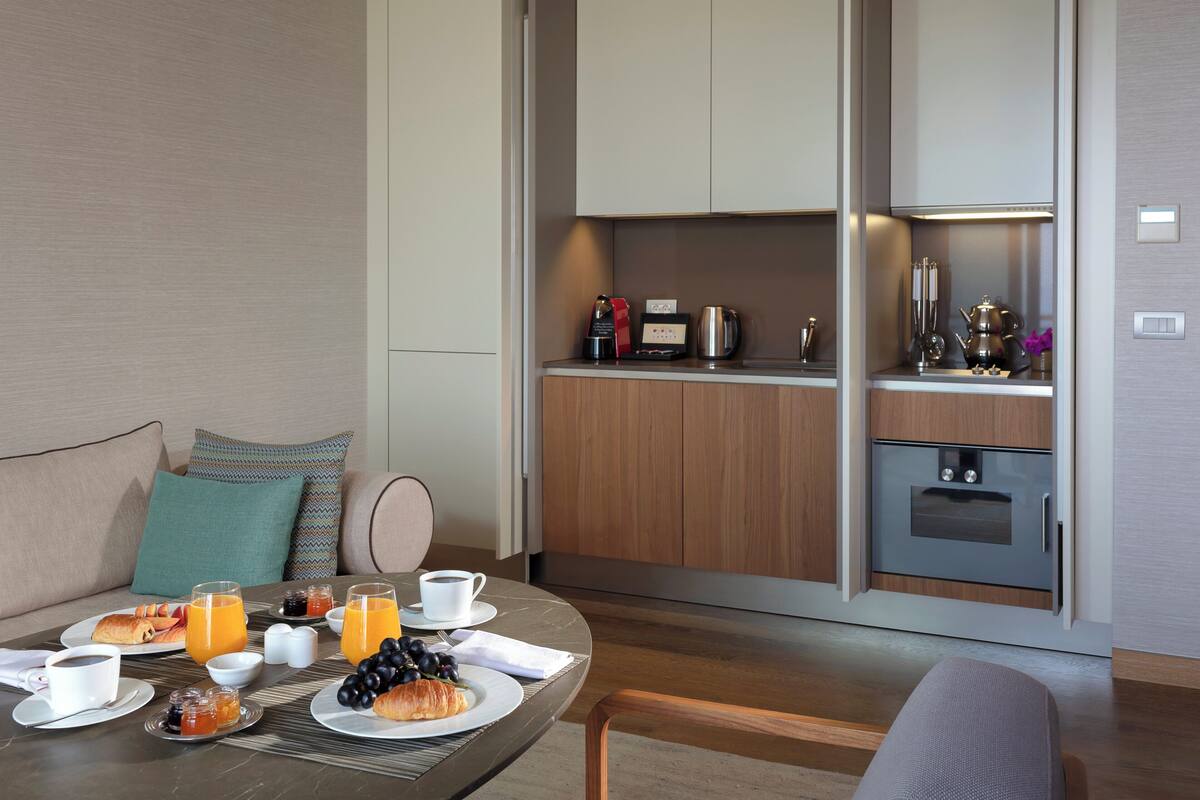 A modern hotel room with a dining area featuring breakfast items, a kitchen with appliances, and a seating area with a sofa and cushions.