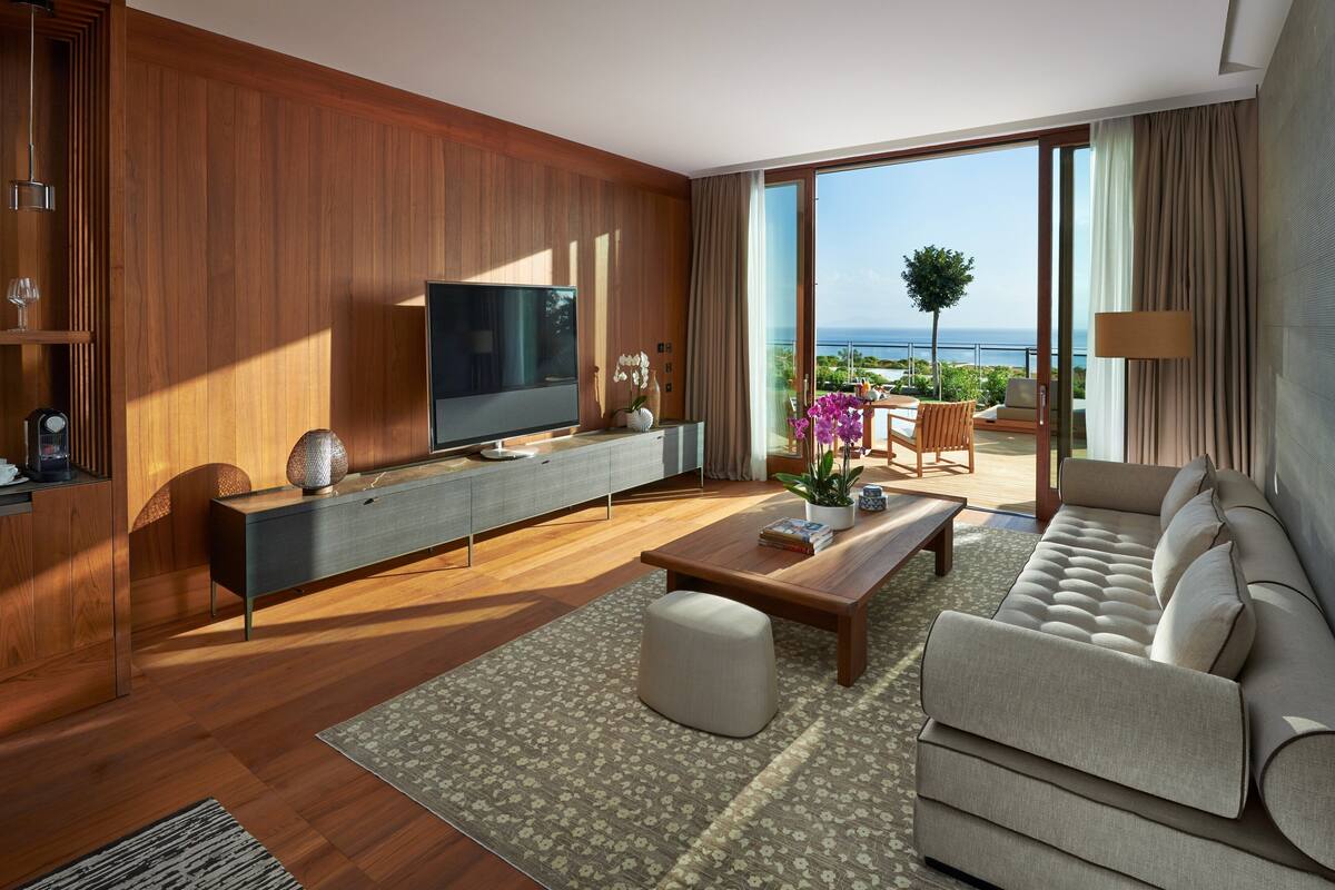 A modern living room with a large flat-screen TV, a sofa, a coffee table, and a balcony with a view of the sea.