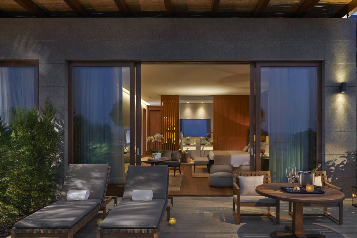 A modern outdoor seating area with lounge chairs and a table, a wooden deck, and a view into the interior of a contemporary home.