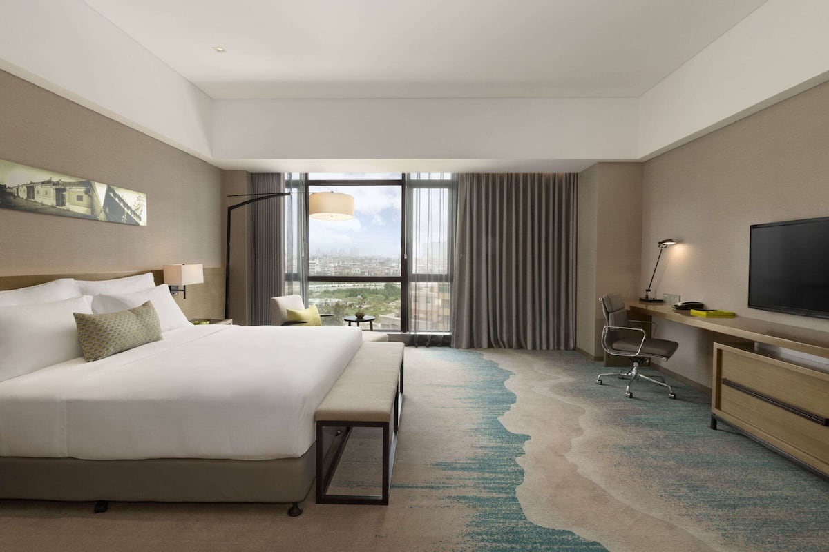 A hotel room with a large bed, a desk with a computer, a TV, and a view of the city through large windows.