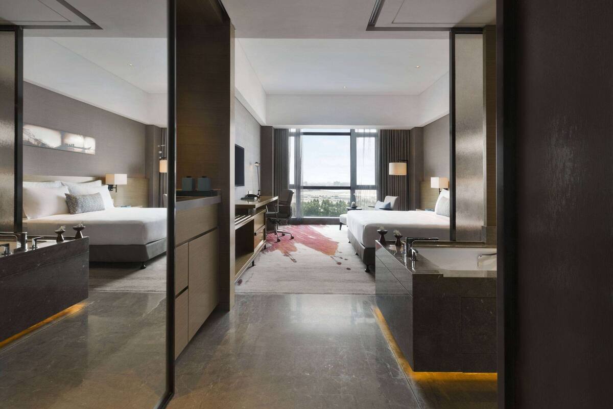 A modern hotel room with two beds, a desk, and a large window offering a city view.