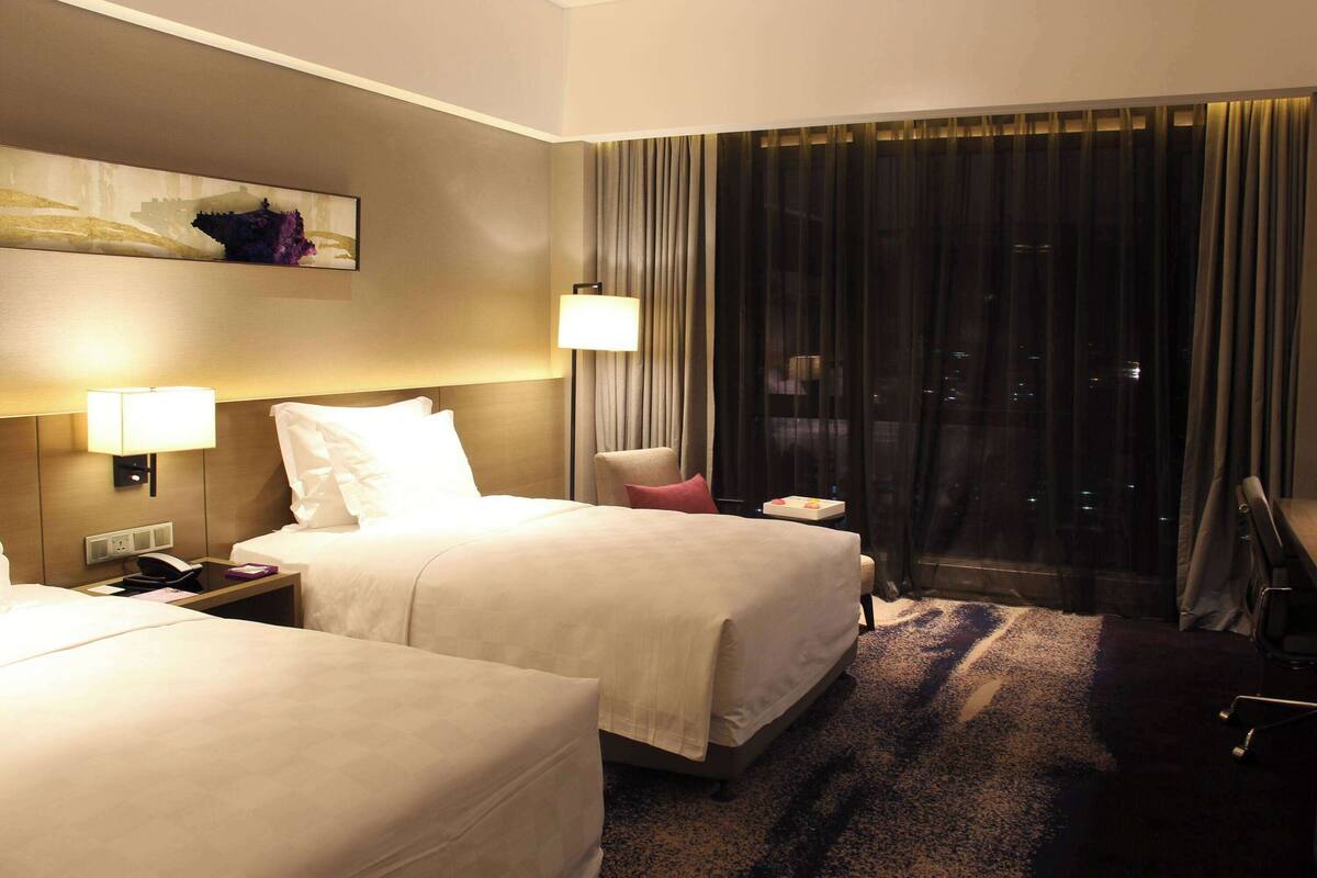 A hotel room with two beds, a nightstand with a lamp, a chair, and a large window with curtains.