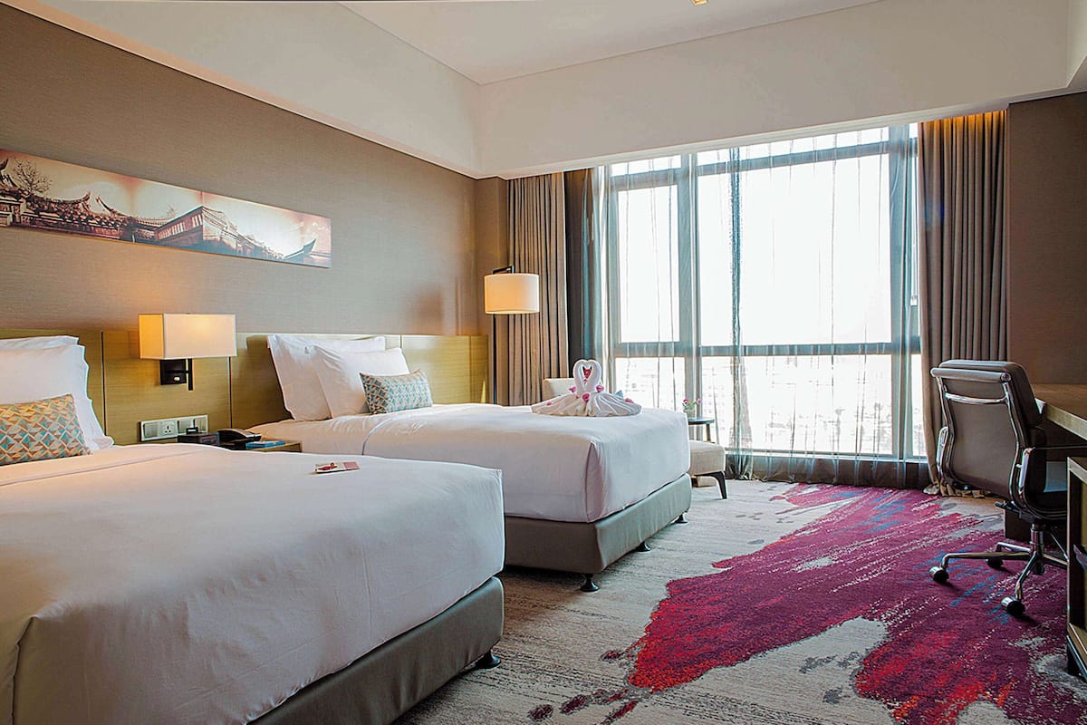 A hotel room with two beds, a desk, a chair, a large window, and a painting on the wall.