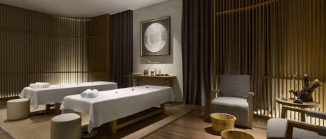 A modern spa room with two massage tables, a wooden floor, and a large wall-mounted artwork.