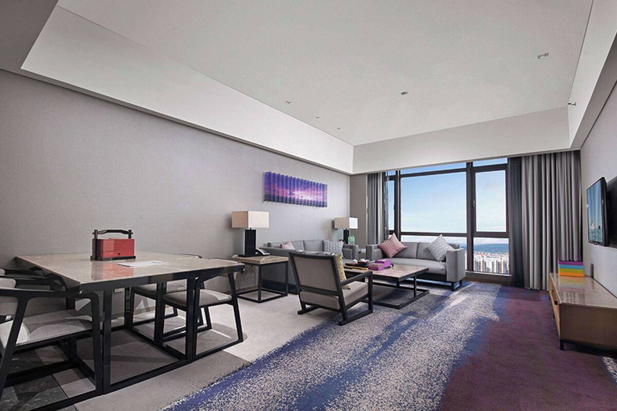 A modern hotel room with a large window, a sofa, a dining table, and a TV.