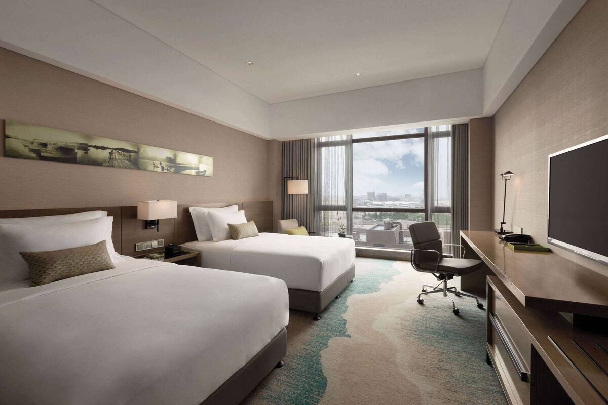 A hotel room with two beds, a desk with a chair, a large window with a city view, and a flat-screen TV.