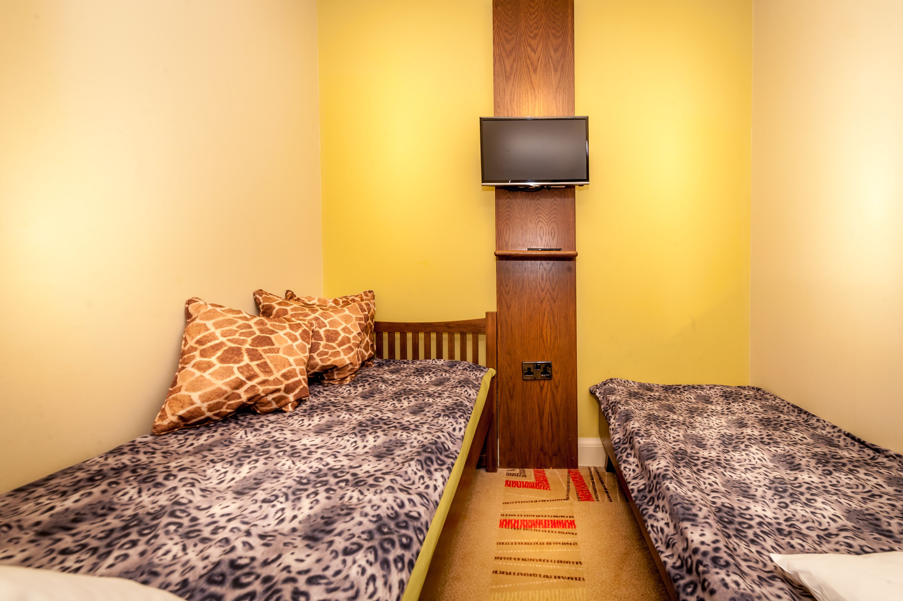 Una habitación pequeña con dos camas, cada una con una colcha de estilo leopardo, un cabecero de madera y un televisor montado en la pared.