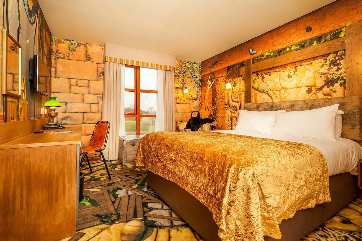 A hotel room with a large bed, a desk, and a chair. There is a window with curtains, a wall with a stone pattern, and a wall with a wooden beam and leaves.