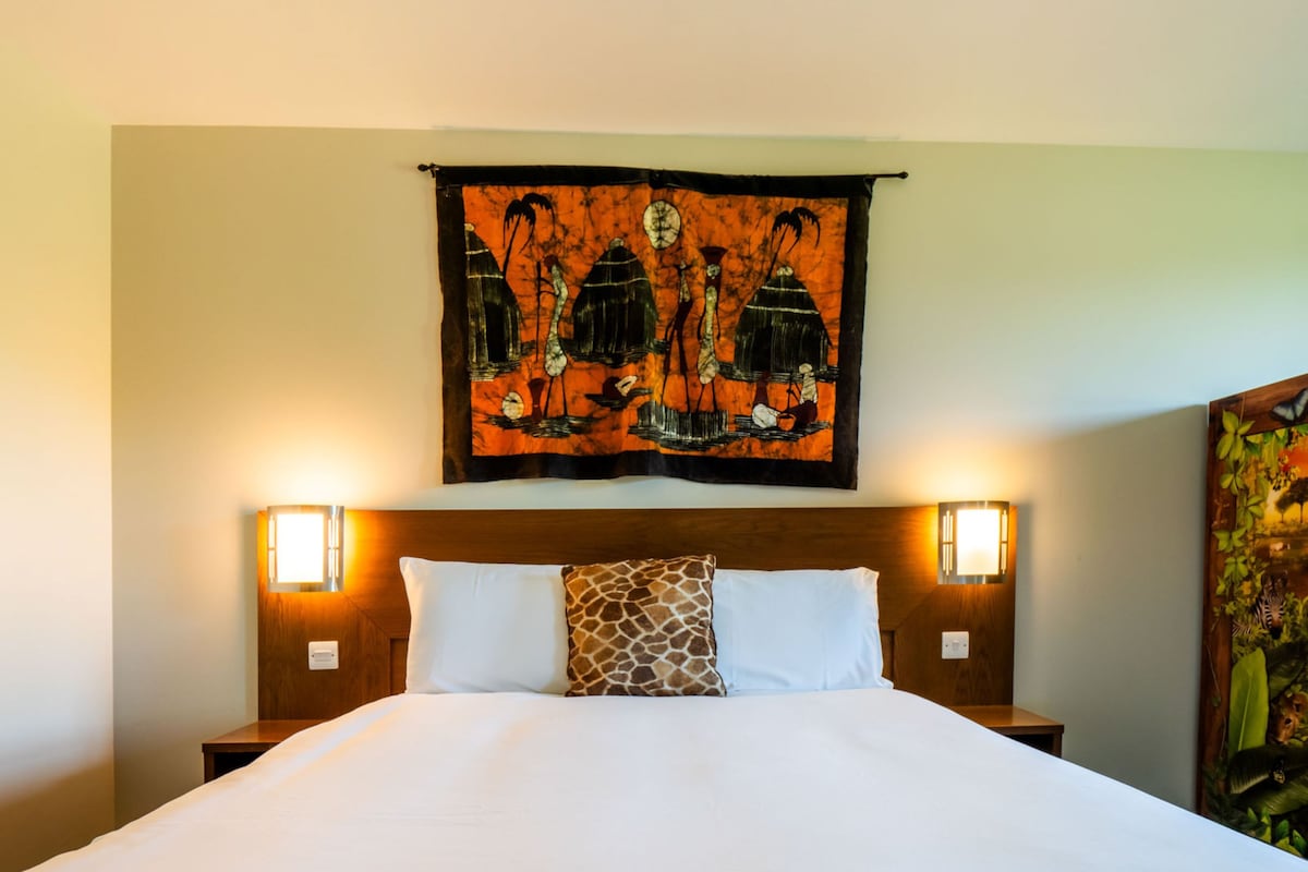 A hotel room with a large bed, two bedside lamps, a headboard with a tapestry, and a wall-mounted artwork.