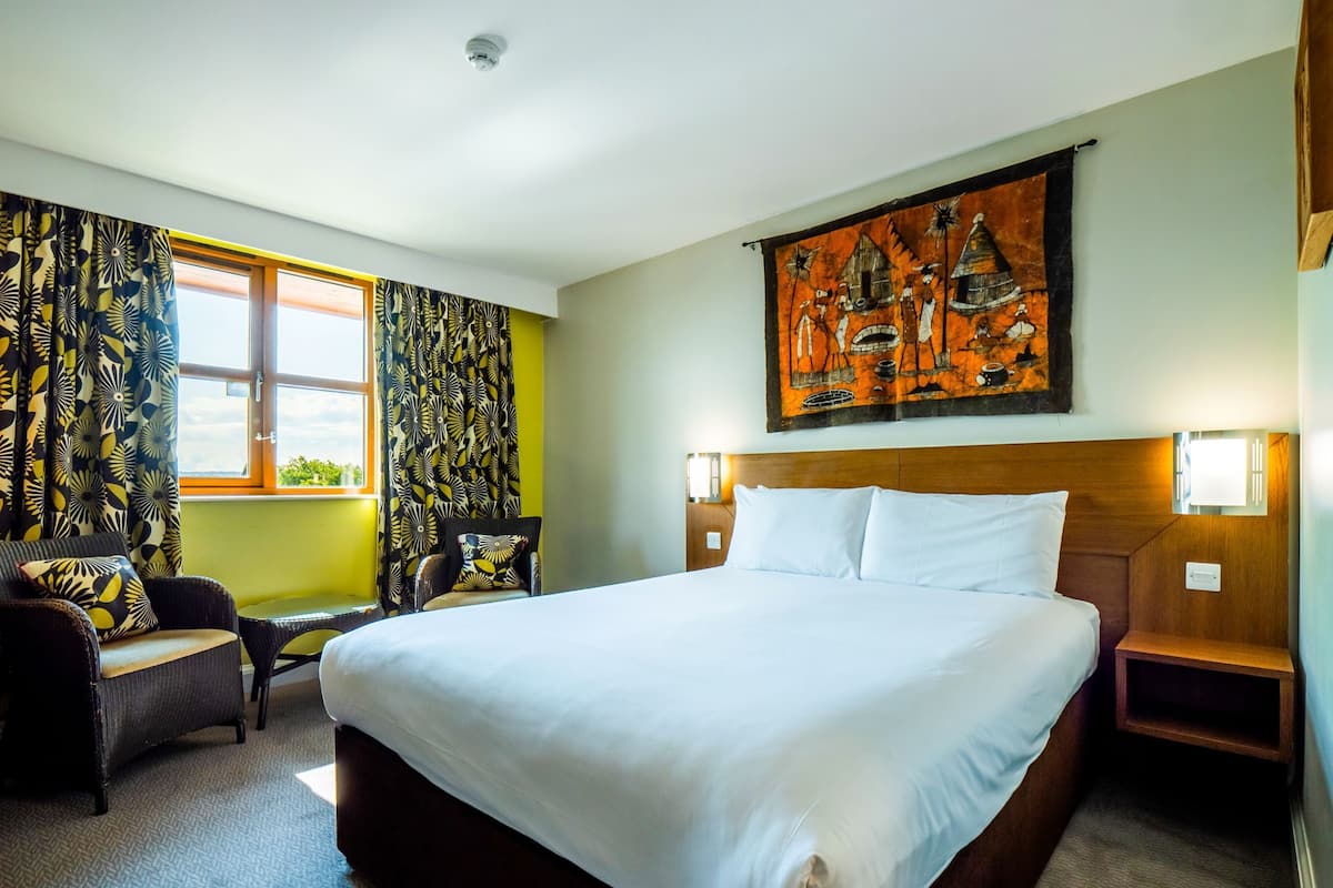 A hotel room with a large bed, a chair, a desk, a window with curtains, and a wall painting.