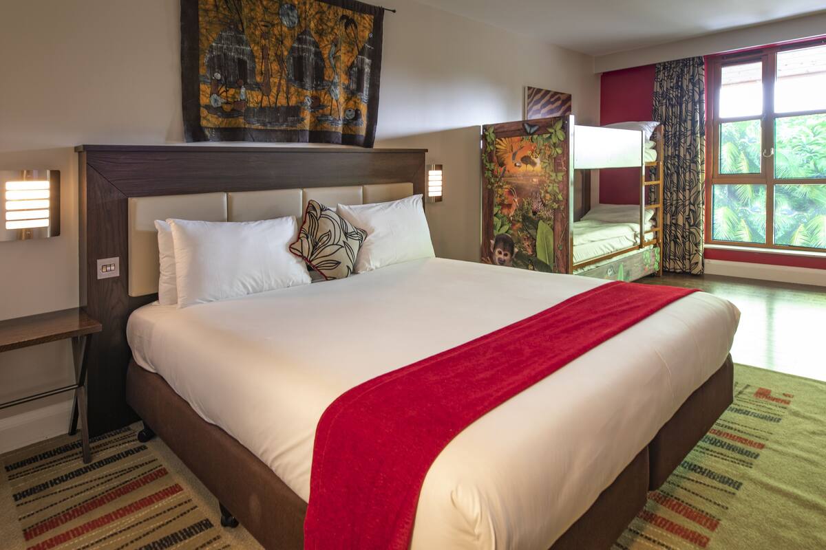 A hotel room with a large bed, a headboard, a nightstand, a wall-mounted lamp, and a window with curtains.