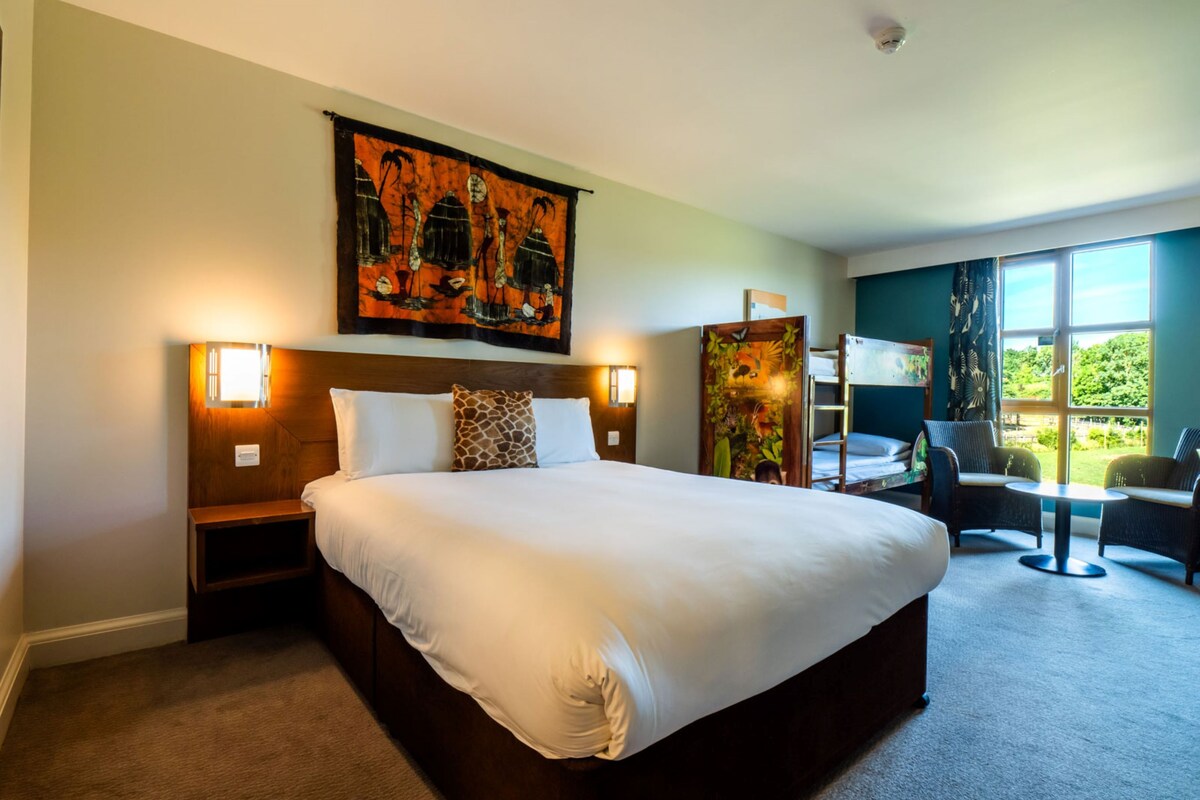 A hotel room with a large bed, bedside lamps, a desk with chairs, a window with a view, and a painting on the wall.