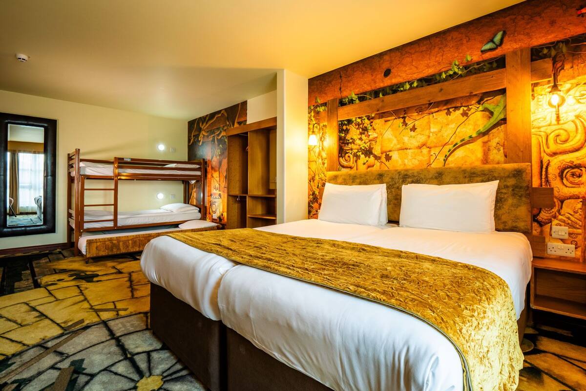 A hotel room with a large bed, a smaller bunk bed, a wooden headboard with a mural, and a patterned floor.