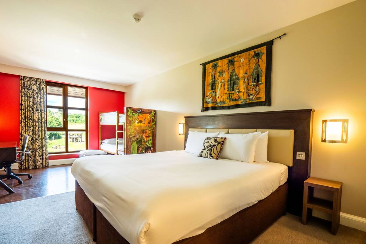 A hotel room with a large bed, a desk, a chair, a window with curtains, and a painting on the wall.