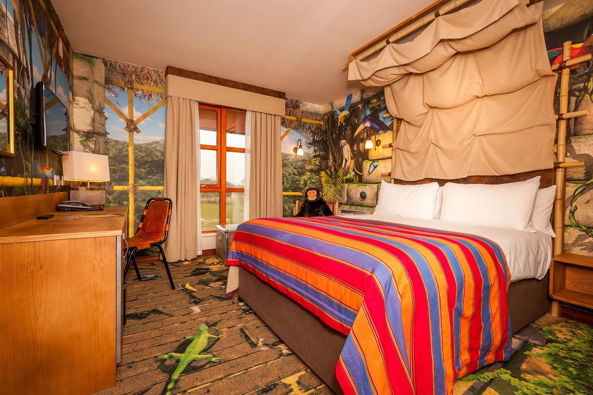 A hotel room with a large bed, a desk, and a chair. There is a window with curtains, a painting on the wall, and a canopy over the bed.
