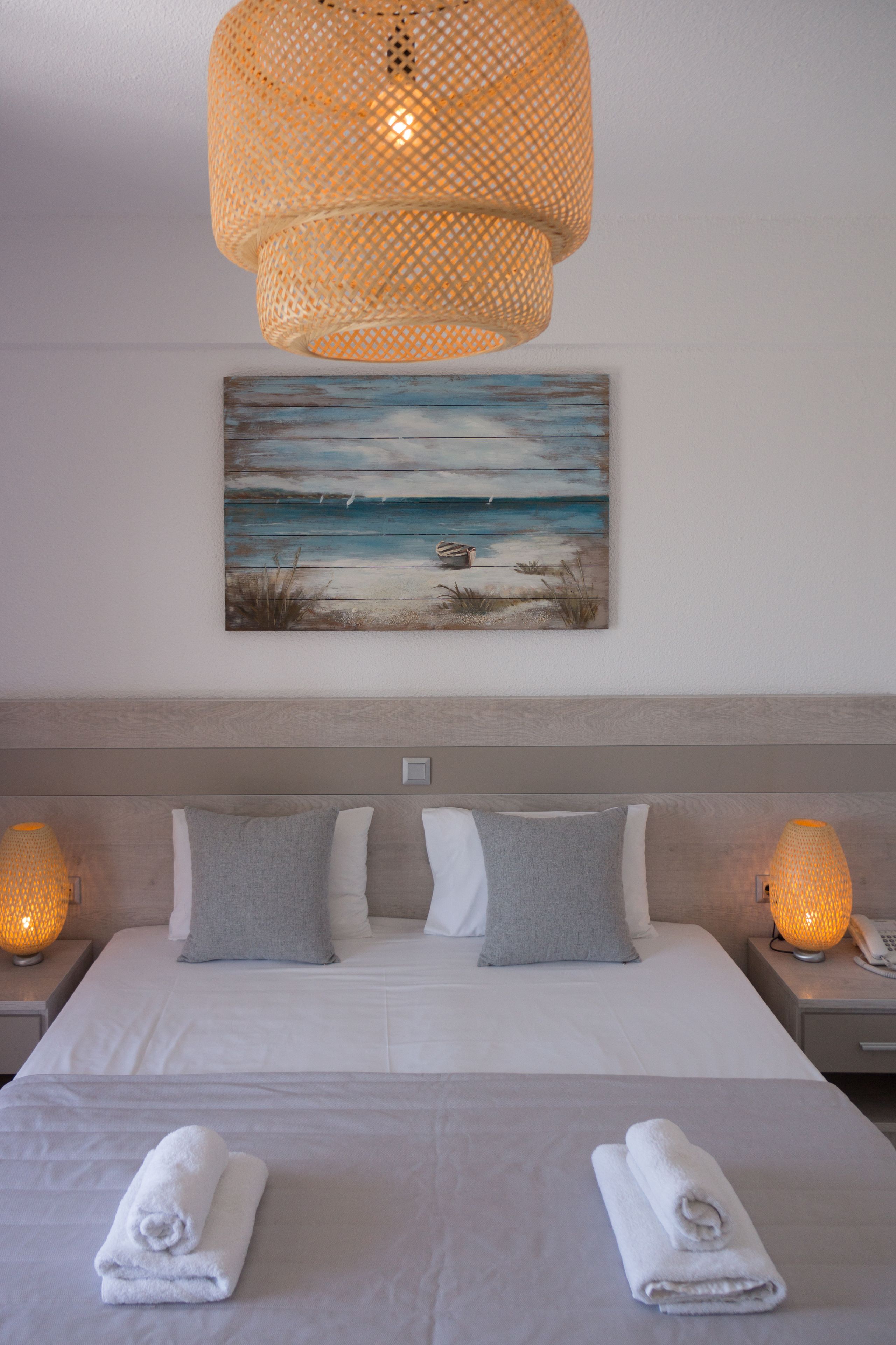 Una cama bien hecha con dos almohadas, un cuadro de una playa y una lámpara de fibra trenzada arriba.