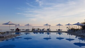 Un área junto a la piscina con sillones de descanso y sombrillas, un cielo despejado y un mar tranquilo al fondo.