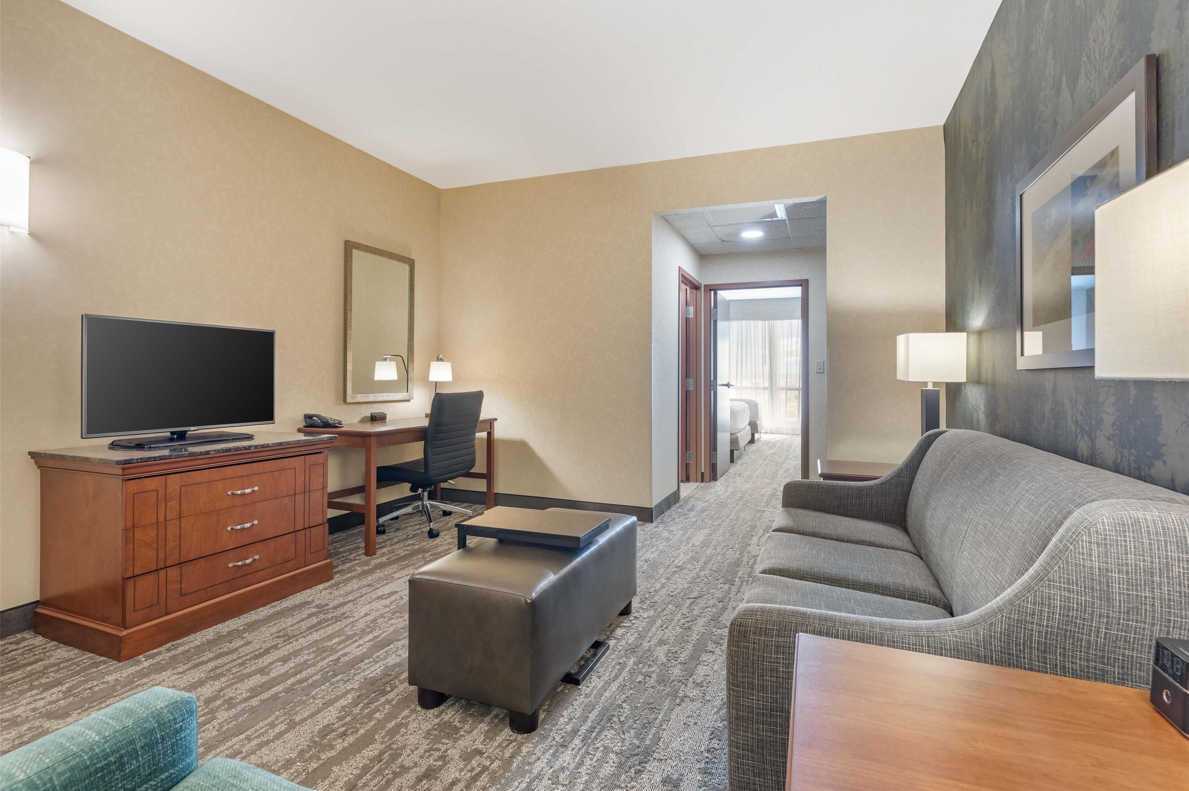A hotel room with a flat-screen TV, wooden dresser, grey sofa, ottoman, and a desk with a chair.