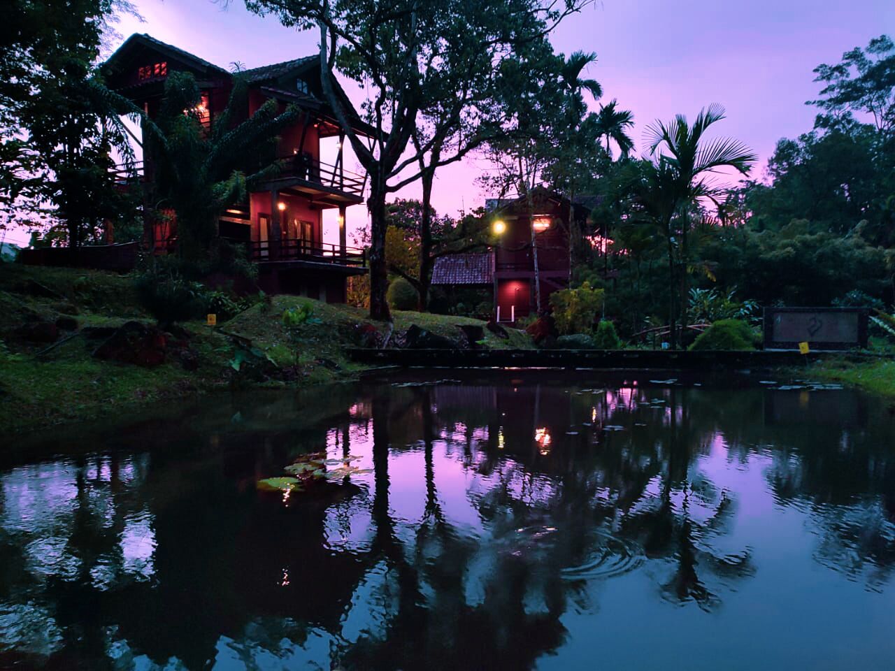 A two-story lodge with a pond in front, surrounded by lush greenery.