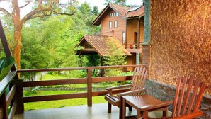 A wooden deck with two chairs and a table, overlooking a lush green garden and a wooden house with a sloped roof. - Sentosa Villa (Taiping)