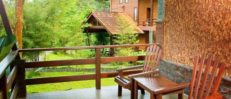A wooden deck with two chairs and a table, overlooking a lush green garden and a wooden house with a sloped roof.