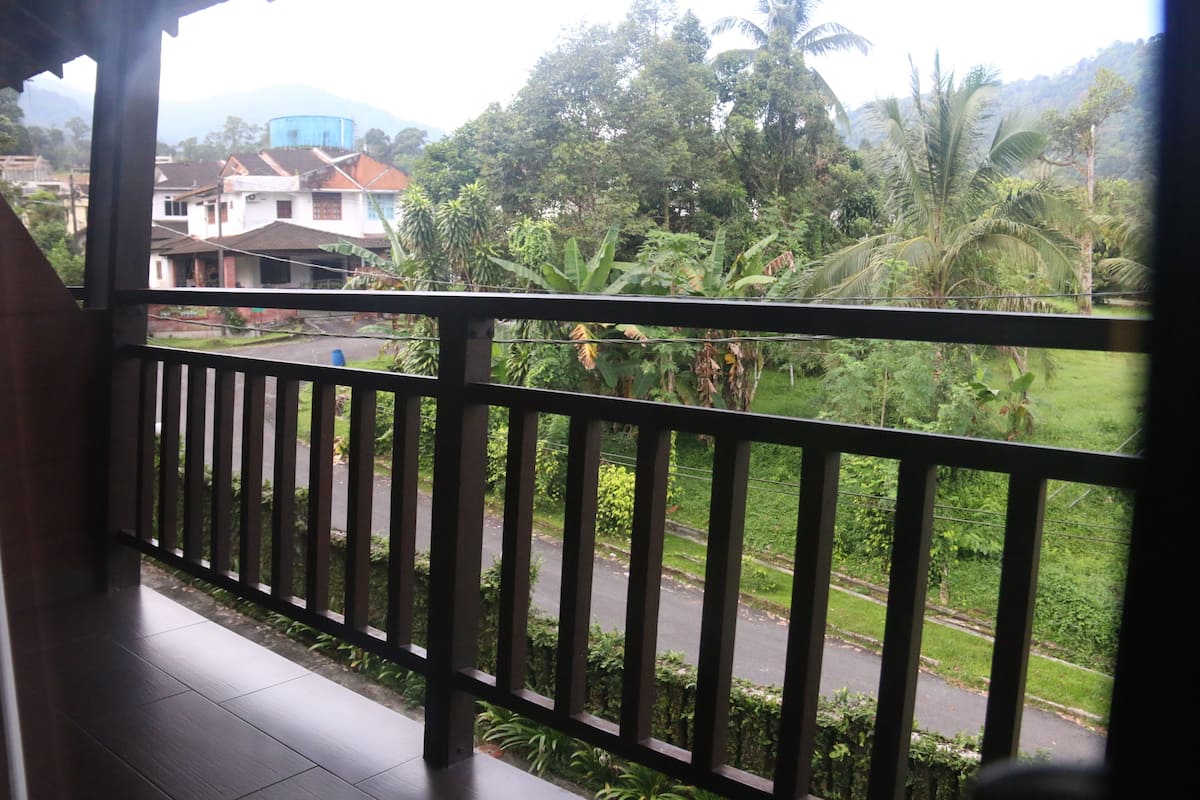 A balcony with a view of a road and greenery, including banana trees and a water tower in the background.