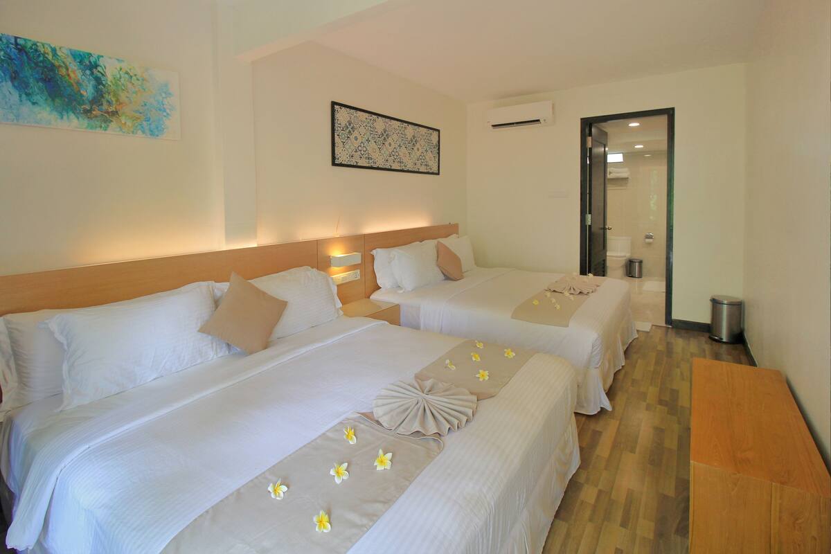 A hotel room with two beds, a wooden bench, a trash bin, and a bathroom visible through an open door.