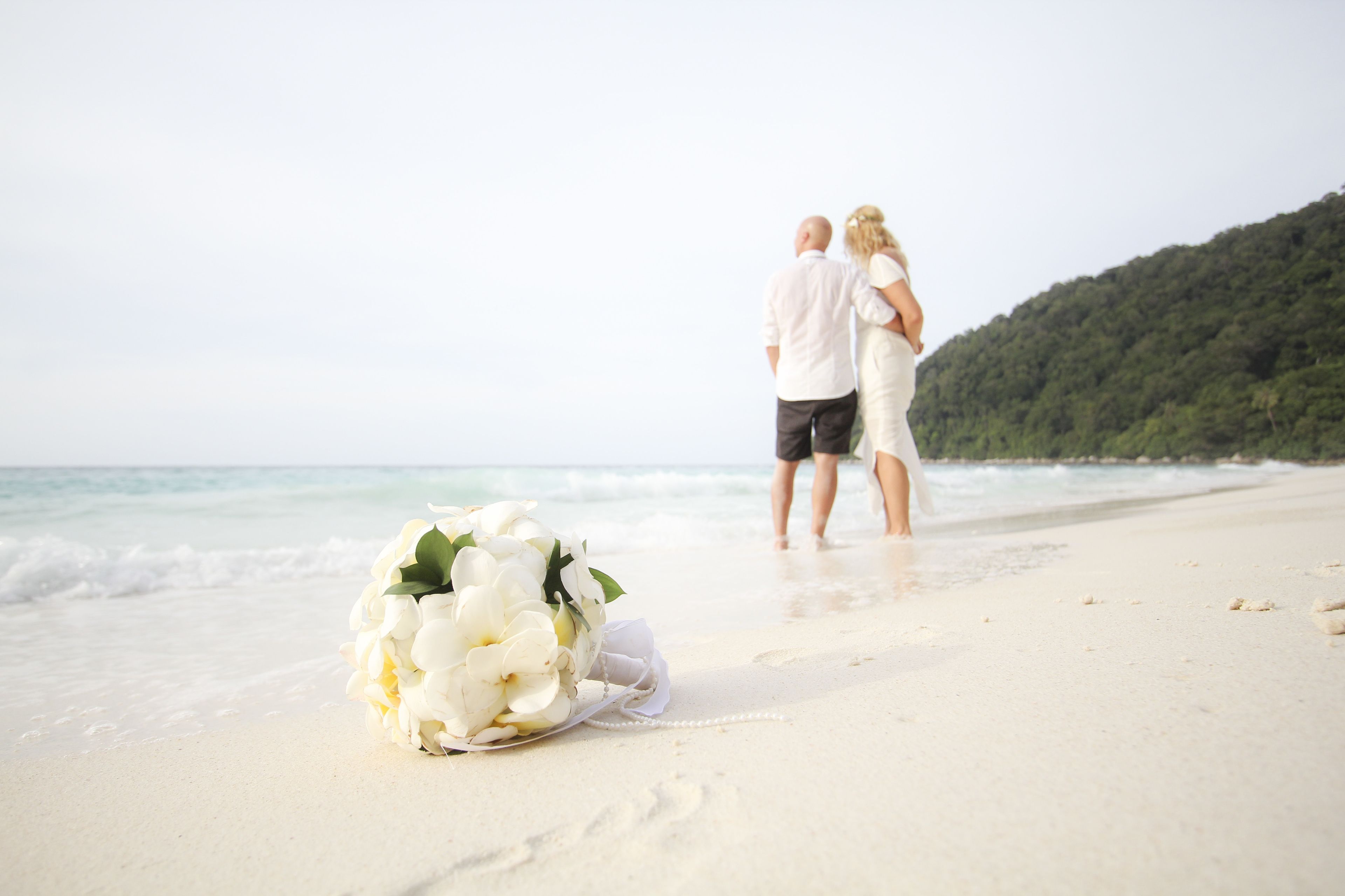 A beach scene with a bouquet of white flowers on the sand, a couple walking along the shore, and a forested hill in the background.