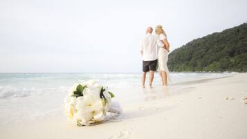 A beach scene with a bouquet of white flowers on the sand, a couple walking along the shore, and a forested hill in the background.